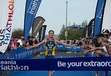 A triumphant male triathlete crosses the finish line at Oceania Triathlon event, surrounded by cheering spectators and vibrant banners, showcasing athletic achievement and endurance.