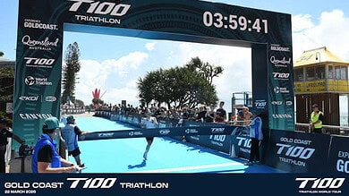 Finish line at the Gold Coast 7100 Triathlon with athletes crossing the water finish, race clock showing 3:59:41, and event branding visible.