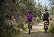 Two cyclists riding mountain bikes on a forest trail during daytime, surrounded by lush green trees and nature.