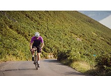 An experienced cyclist wearing a purple jersey and helmet riding along a winding mountain road surrounded by lush greenery and hills, showcasing outdoor endurance training.