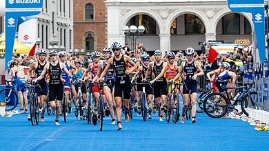 Triathlon athletes start race in urban city setting, walking with bikes on blue carpet, with spectators and historic buildings in background.