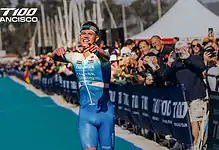 A high-energy moment at the San Francisco triathlon, featuring a smiling athlete crossing the finish line in a blue triathlon suit, surrounded by cheering spectators and event banners.